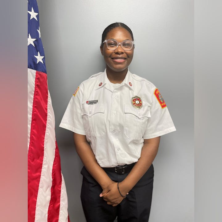 A person in a fire department uniform stands beside an American flag, smiling.