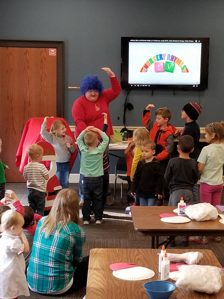A group of children participating in an activity led by an adult with a blue wig; a TV screen displays "NURSERY RHYMES" in the background.
