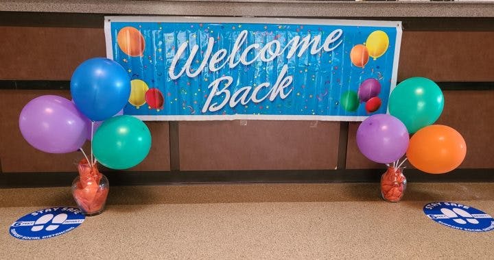 A "Welcome Back" banner with colorful balloons tied to weights, over a floor sign shaped like a paw print.