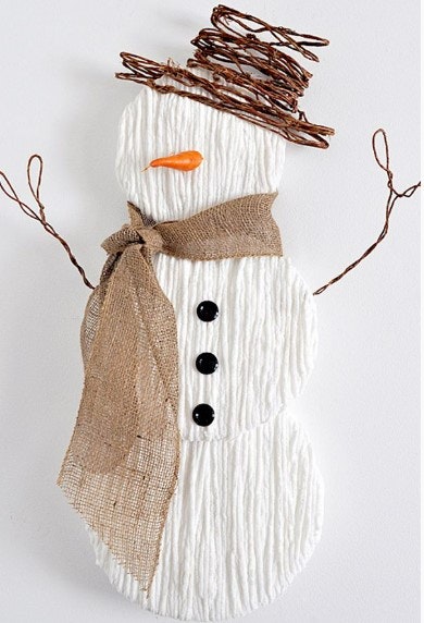 A craft snowman made from corrugated cardboard, with a burlap scarf, twig hat and arms, carrot nose, and button eyes.