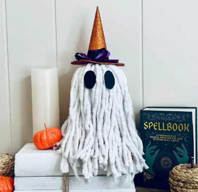 A cute ghost decoration with a witch hat, surrounded by a pumpkin, candle, books, and spellbook, evoking a Halloween theme.