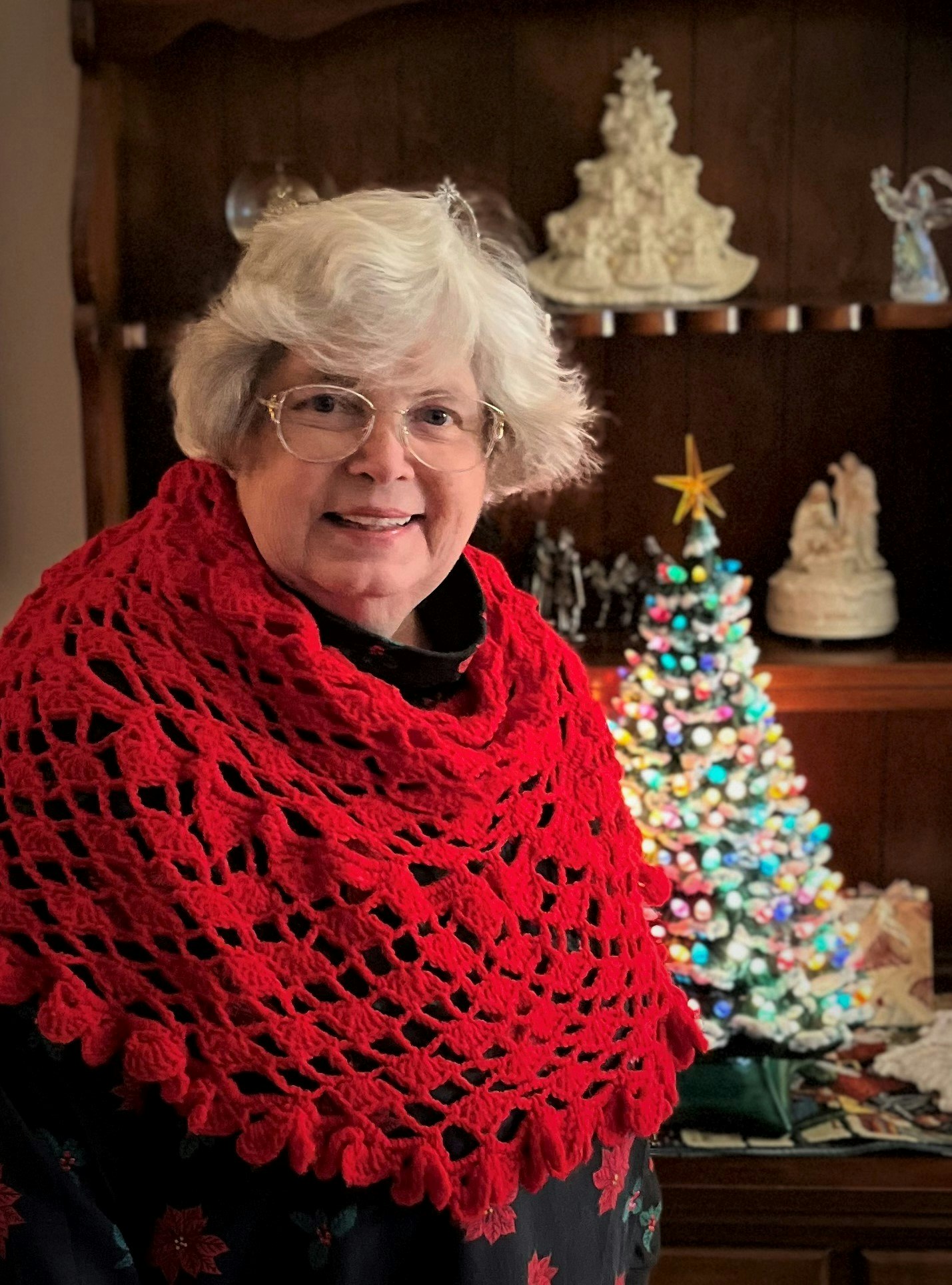 A person with a red crochet shawl in front of a decorated Christmas tree and holiday decorations.