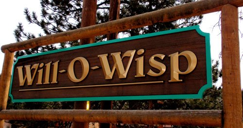Wooden sign with "Will-O-Wisp" in gold letters, surrounded by trees and wood posts.