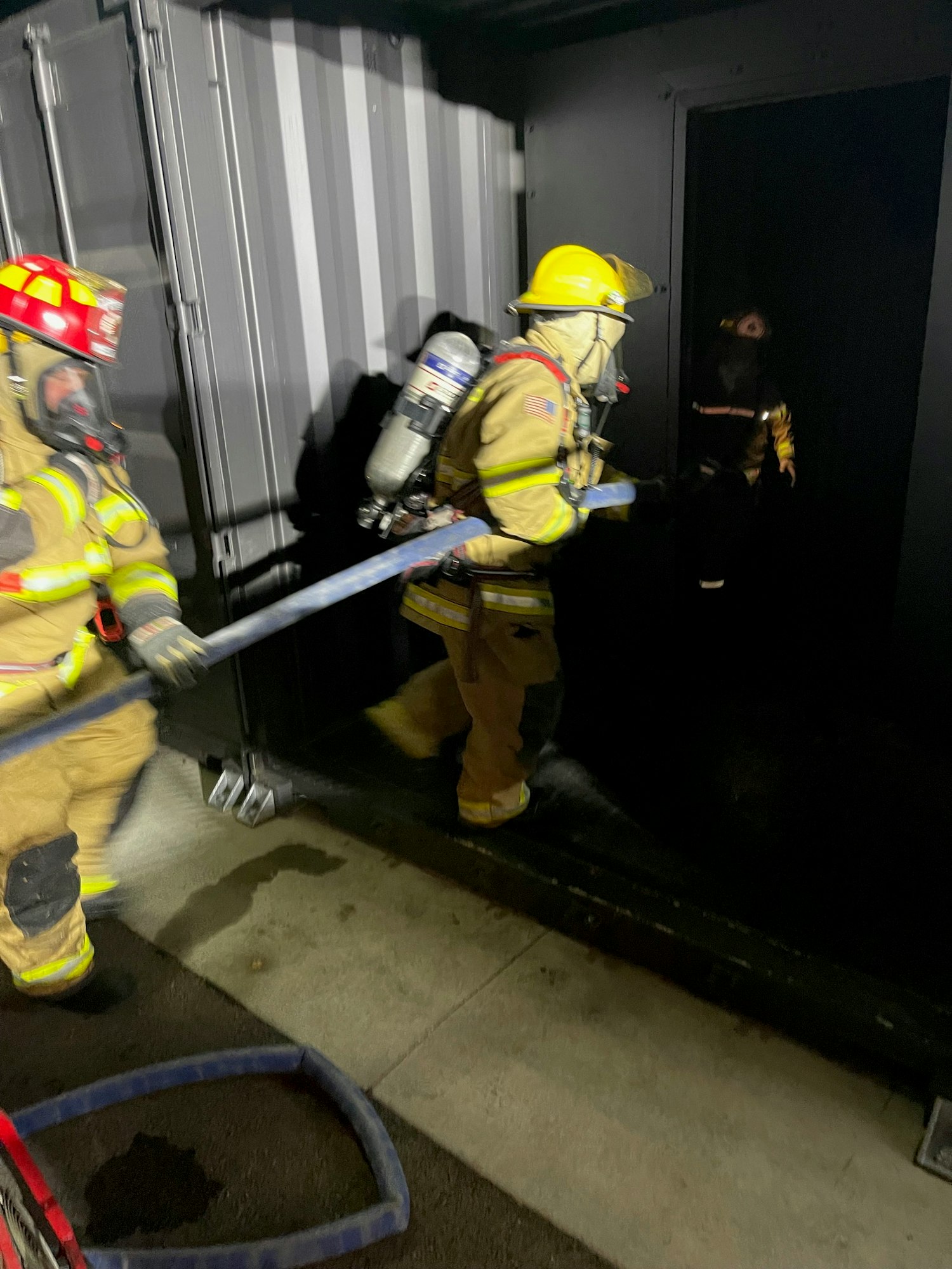 Firefighters in gear performing a task in a dimly lit environment with stairway and hose visible.