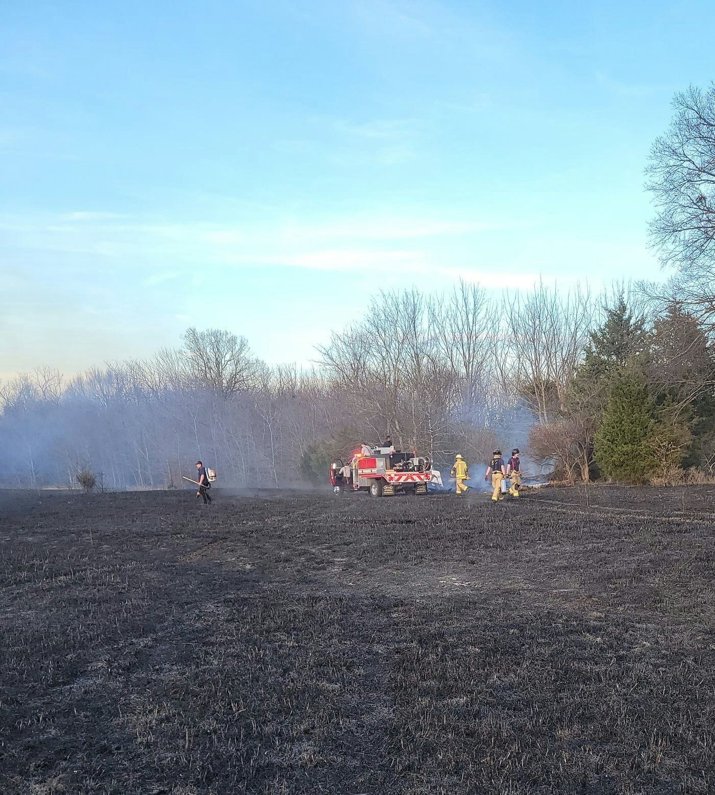 Firefighters assessing a charred landscape with a fire truck, amid lingering smoke and bare trees in the background.