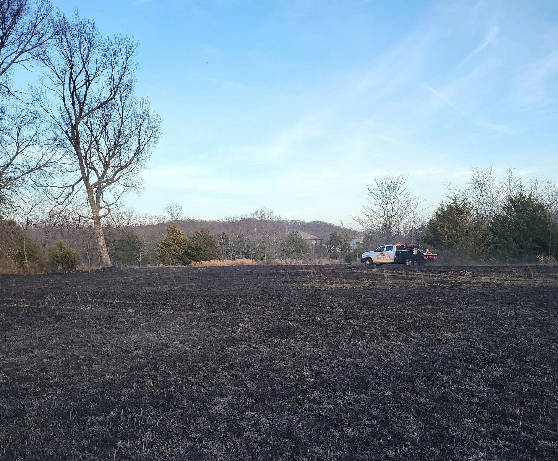 A fire truck parked on a field with burned grass, surrounded by trees and open sky.