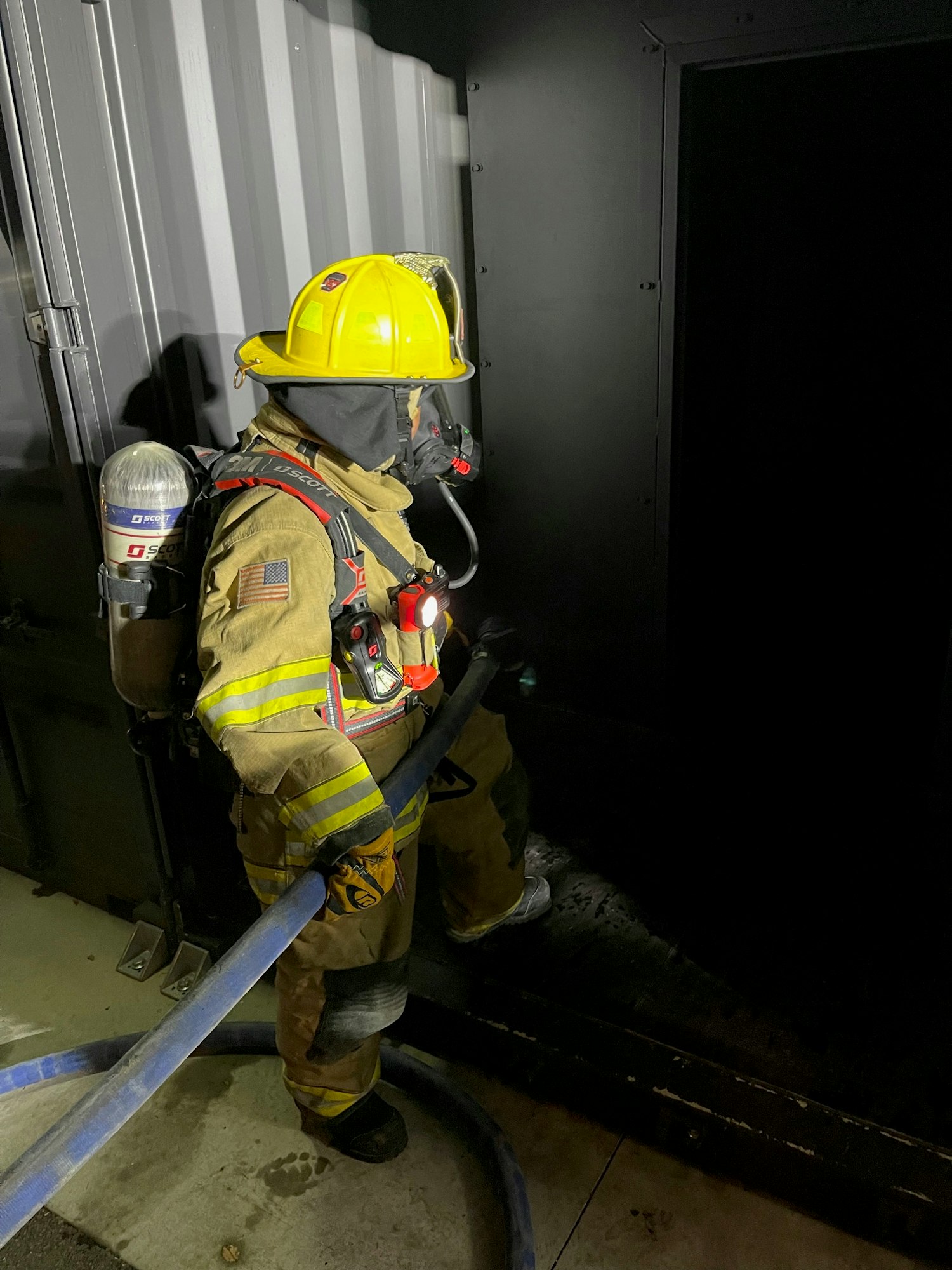 A firefighter in full gear is crouched down, inspecting a structure at night.