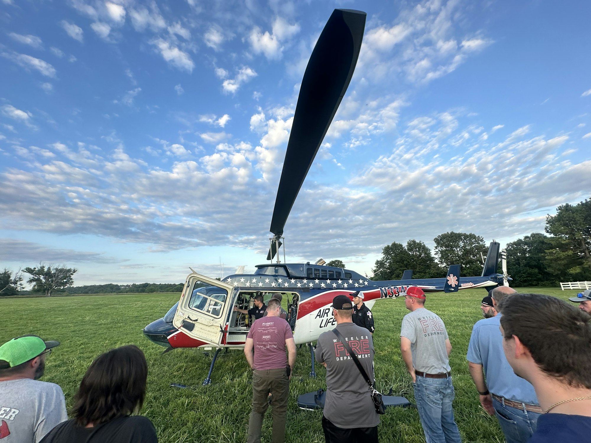A group of people observing a helicopter with a patriotic design on a grassy field under a cloudy sky.