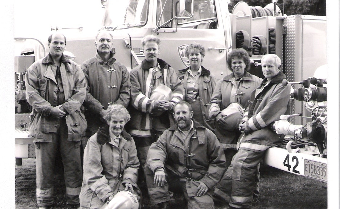 A group of firefighters in uniform poses in front of a fire truck, holding helmets, in a black-and-white photo.
