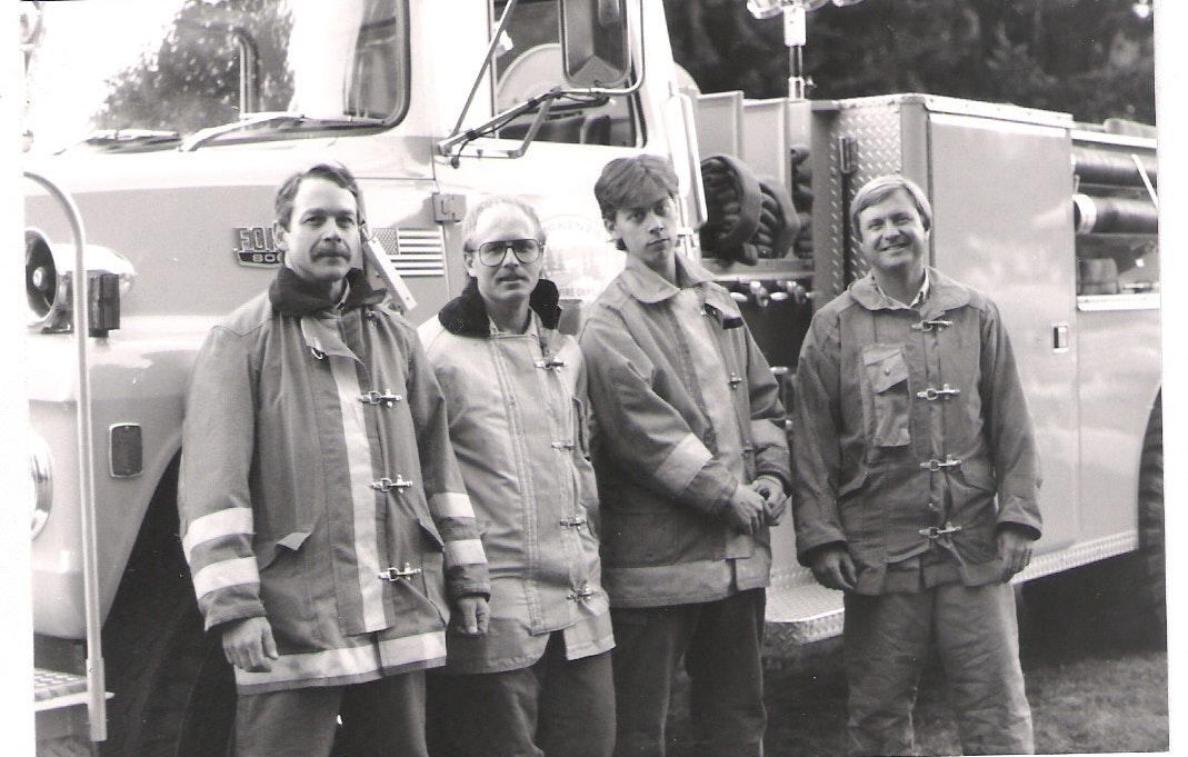 Four people in firefighting gear standing in front of a fire truck. Black and white image.