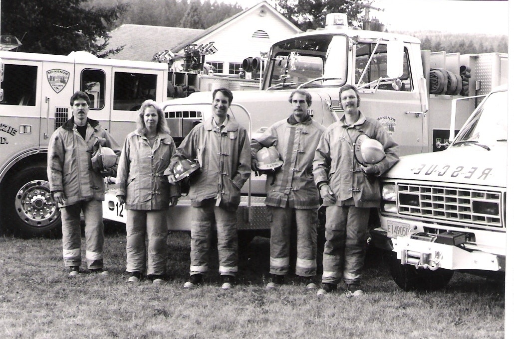 Five firefighters in gear standing in front of fire trucks.