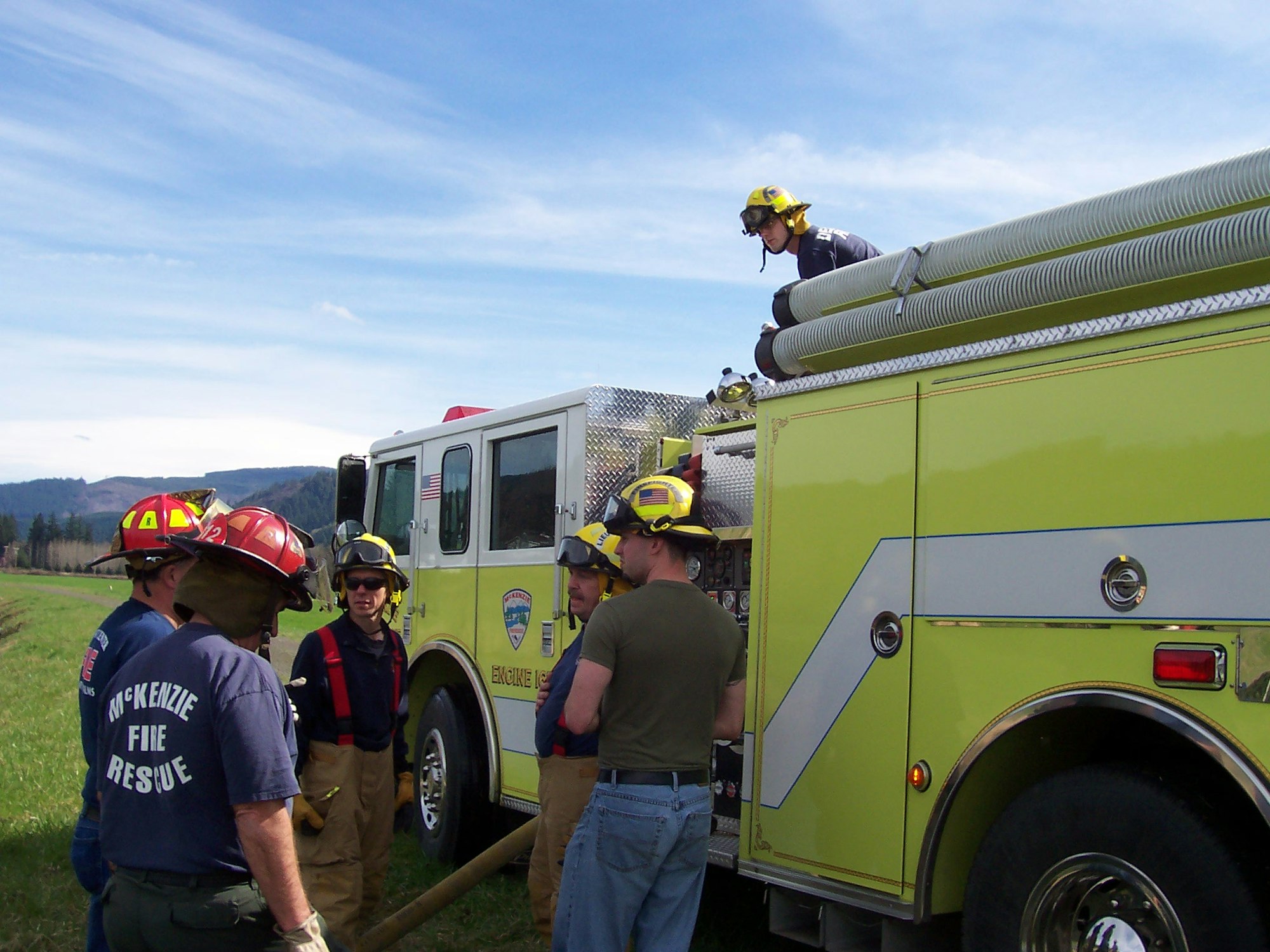 Firefighters in gear stand near a green fire truck, preparing equipment.