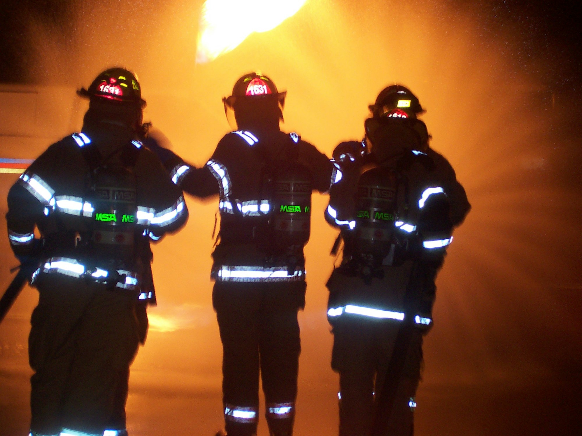 Three firefighters, wearing reflective gear, facing flames in the background.