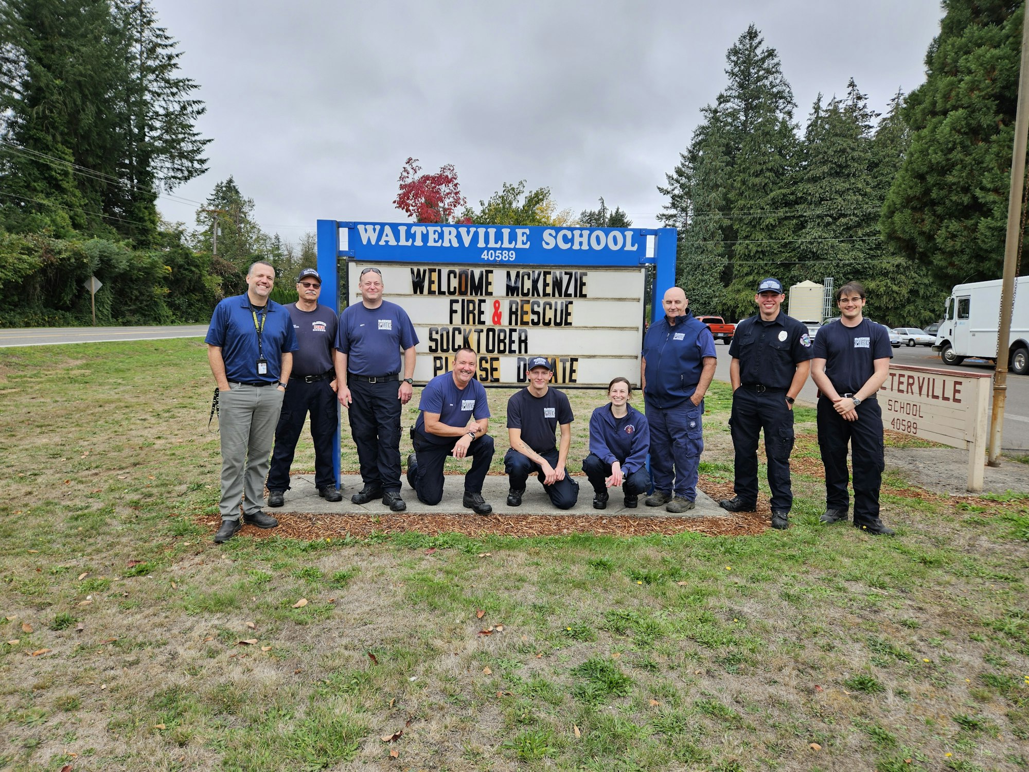 A group of people posing in front of a school sign promoting a fire & rescue event and SOCKtober donations.