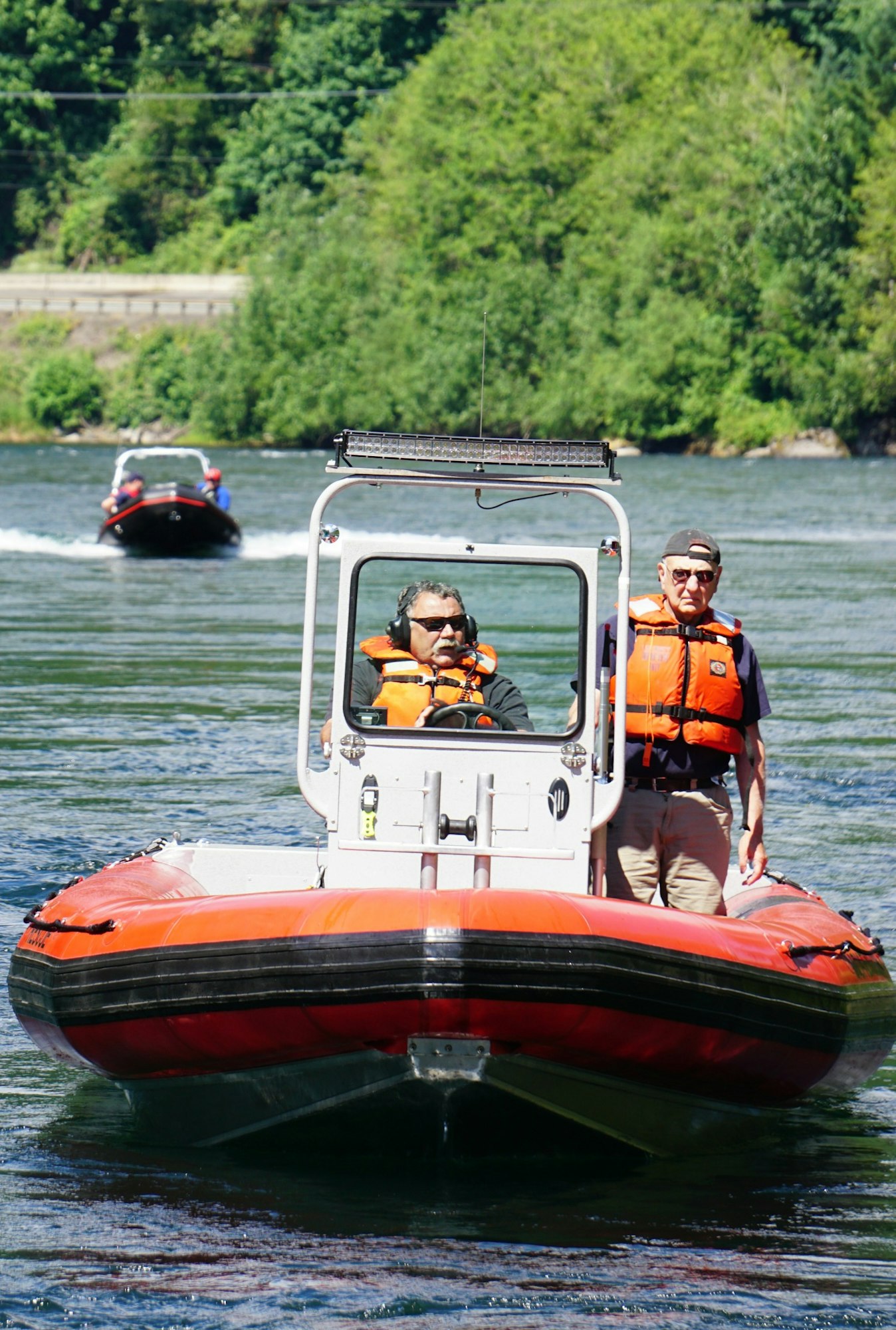Two people in orange life vests on a red boat on a river, with another boat in the background amidst green trees.
