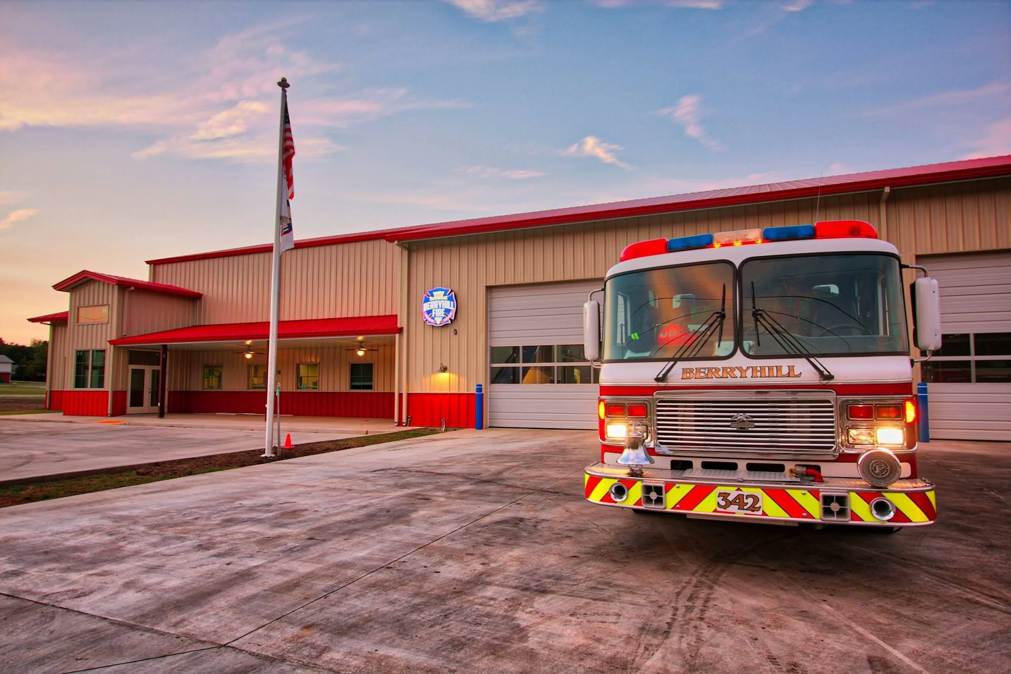 Image of a fire station with a fire truck parked out front, featuring a flag and a colorful sunset backdrop.