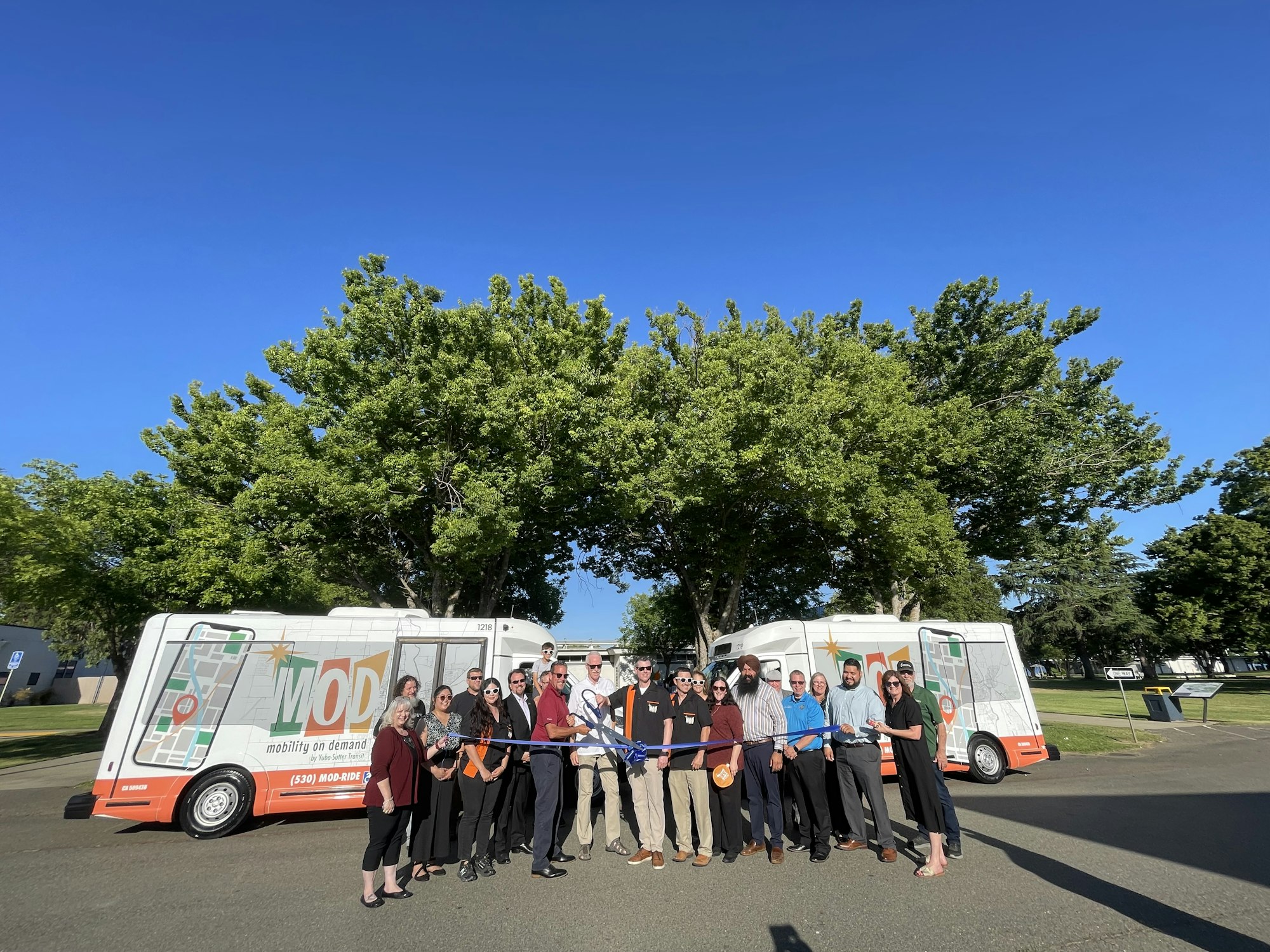 A group of people at a ribbon-cutting ceremony in front of two "MOD" vans under clear blue skies.