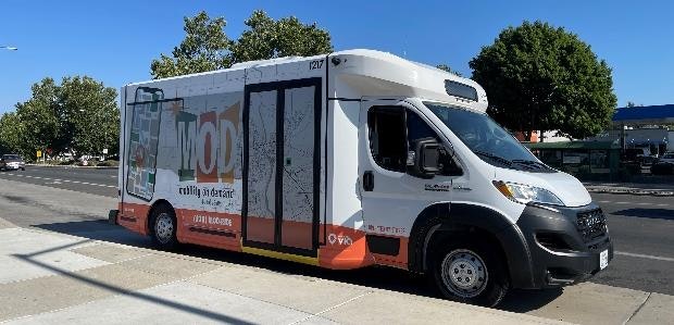 A white and orange mobile library vehicle parked on the street with trees and a sidewalk nearby.