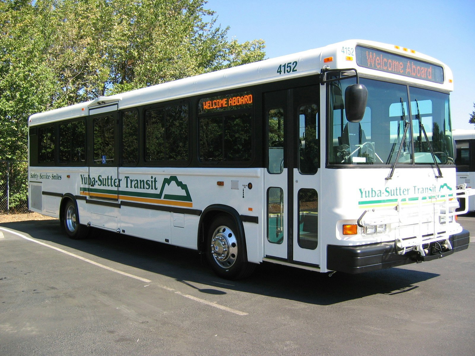 A Yuba-Sutter Transit bus with "Welcome Aboard" on the display, parked near trees on a sunny day.
