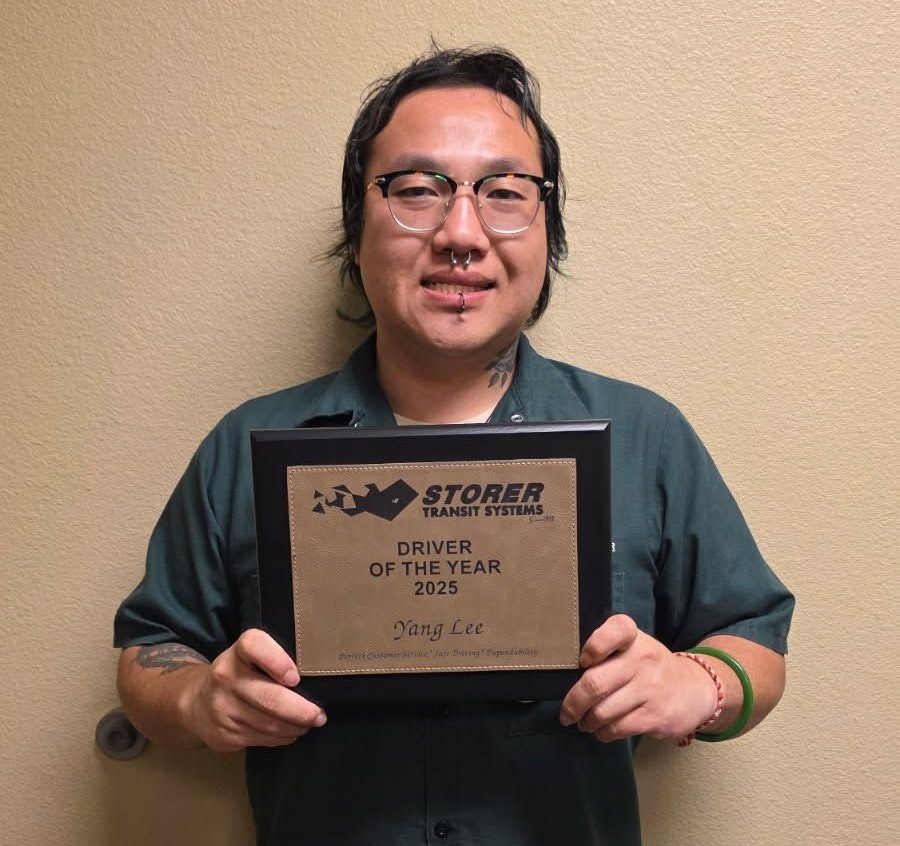 A person holding an award plaque that reads "Driver of the Year 2025" from Storer Transit Systems, named Yang Lee.