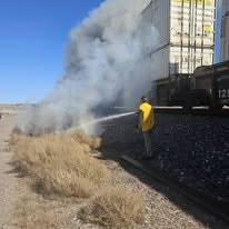 A person in a yellow jacket extinguishes a fire near train tracks, with smoke and dry grass in the foreground.