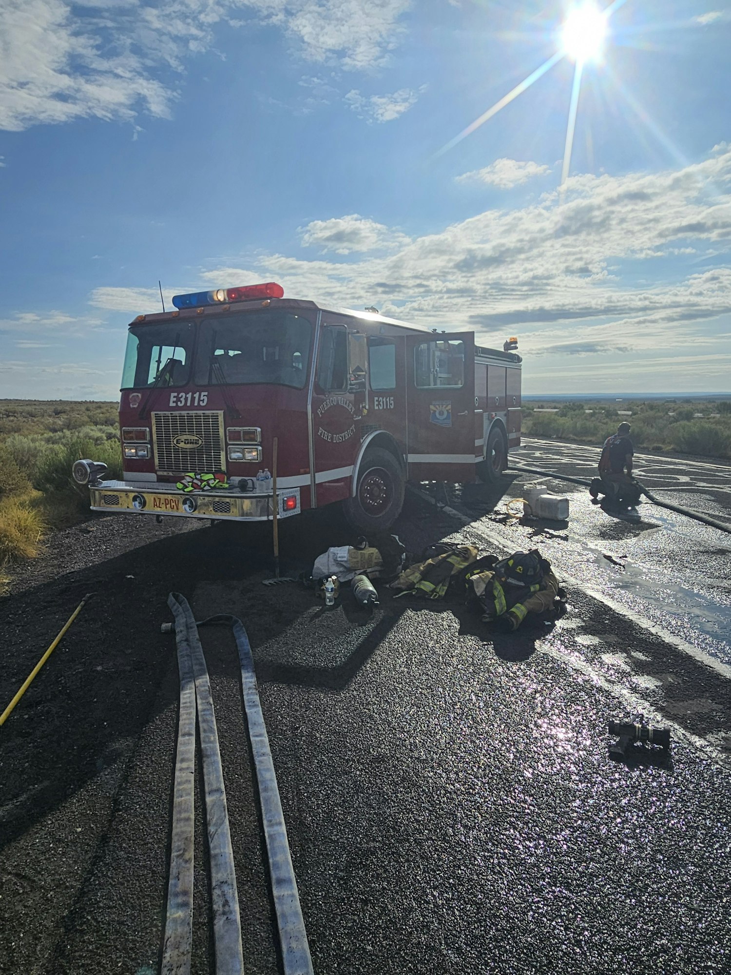 A fire truck on a road with firefighters resting and equipment scattered under a bright sun.