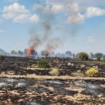 A grassy field on fire with smoke and flames, under a partly cloudy sky.