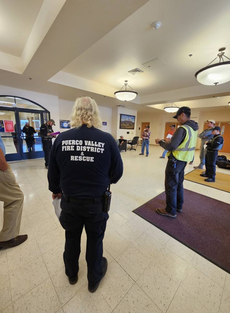 People gathered indoors, one wearing a "Puerco Valley Fire District & Rescue" shirt, appearing to attend a meeting or briefing.