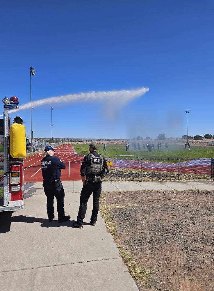 Two security personnel watch a fire truck spraying water towards a field on a clear day.