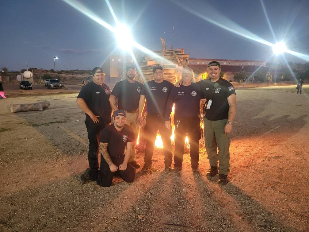Six people in matching uniforms pose in front of a small fire at night, with bright lights in the background.