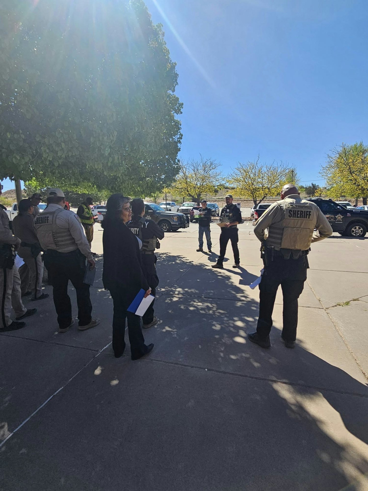 A group of law enforcement personnel gathered outside, some in sheriff vests, with police vehicles in the background.