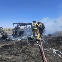 Firefighters extinguishing a smoldering, charred vehicle in a field with a fire hose.