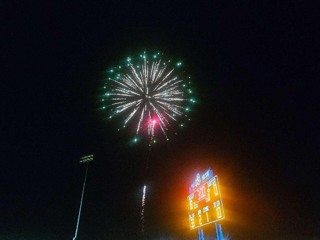 Fireworks burst in the night sky above a lit sports scoreboard.