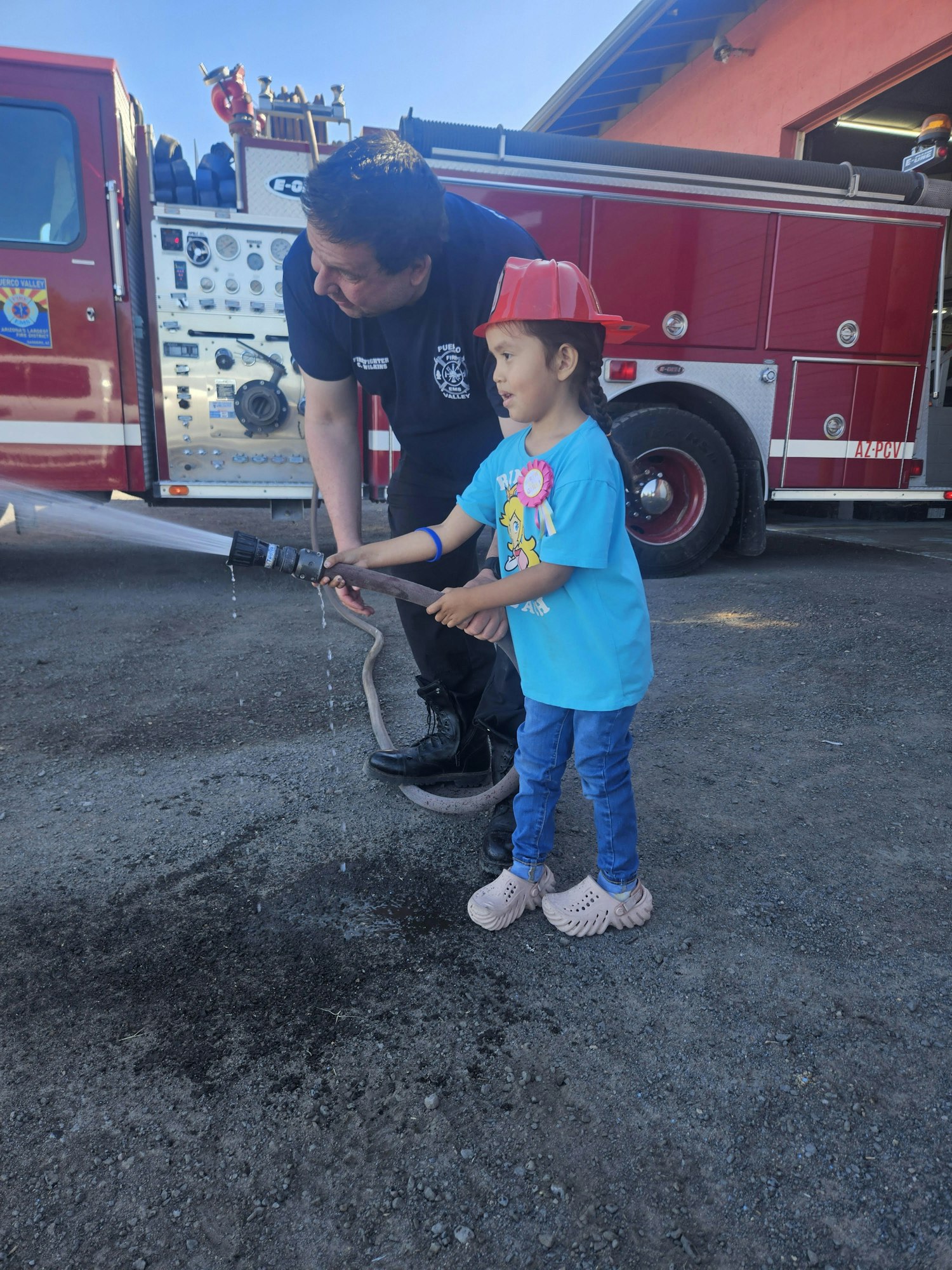 A child wearing a red firefighter hat sprays water from a hose, assisted by an adult in front of a fire truck.