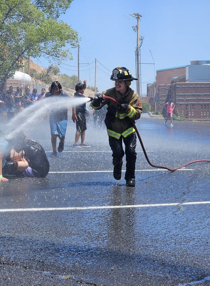 A firefighter sprays water from a hose, splashing people on a sunny day.