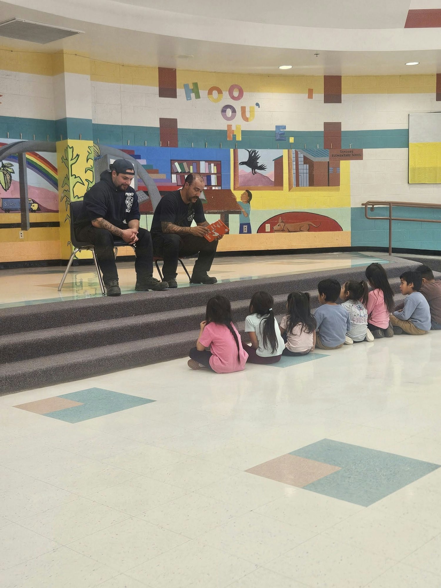 Two people reading to a group of kids sitting on the floor in a colorful room.