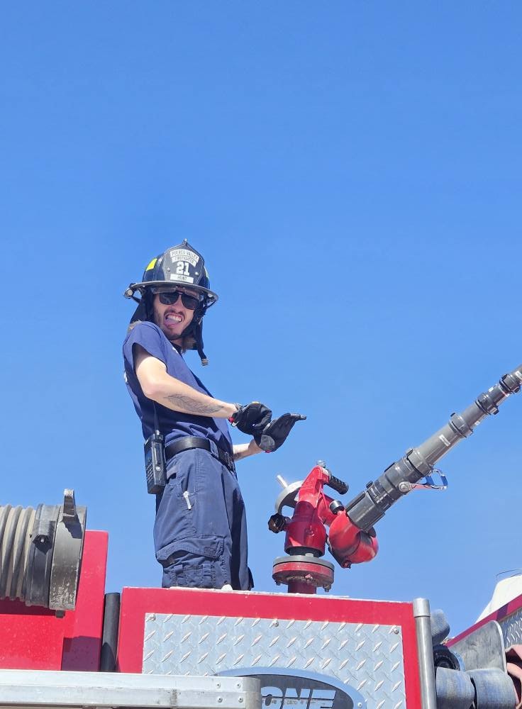 A firefighter in uniform on a fire truck, standing beside equipment, smiling under a clear blue sky.
