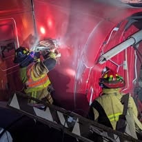 Firefighters using tools on a red vehicle, with smoke around them.