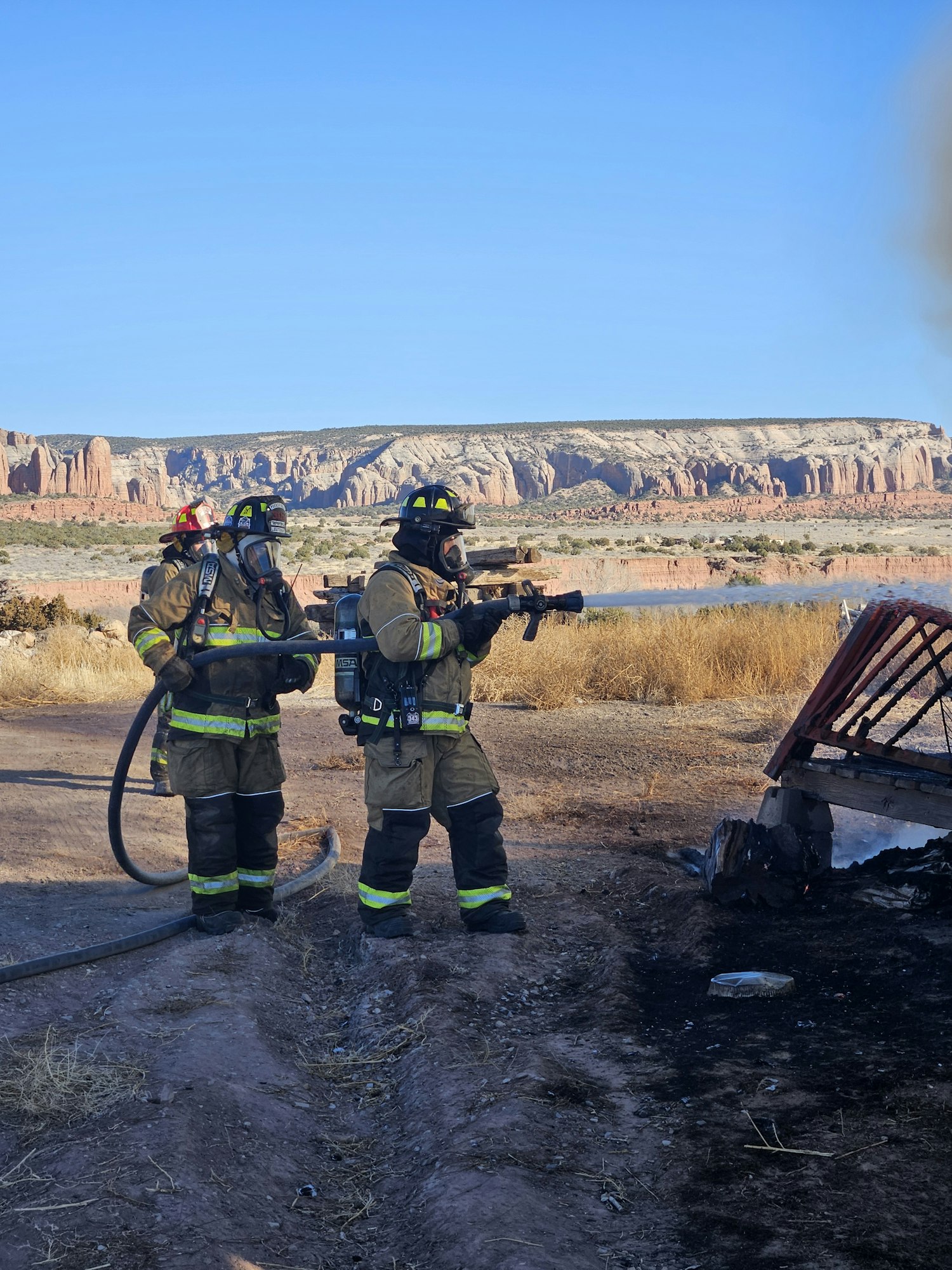 Two firefighters in gear, holding a hose, near a burned area with cliffs and clear blue sky in the background.