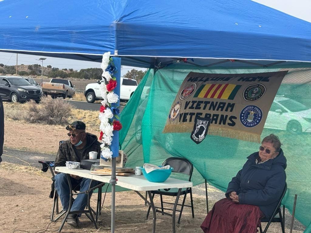 Two people sitting under a tent with a "Welcome Home Vietnam Veterans" banner, with military logos and flowers.
