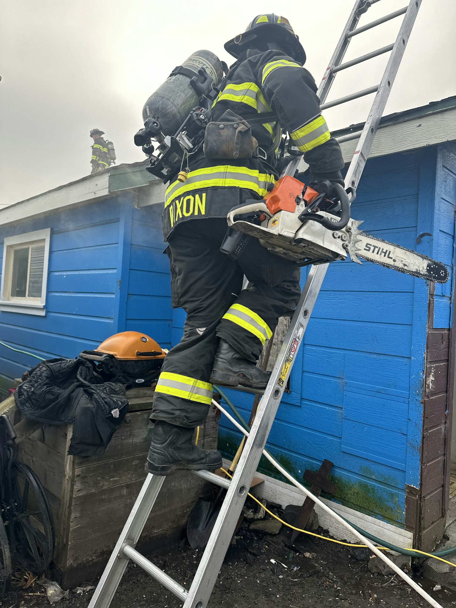 A firefighter with a chainsaw on a ladder by a blue building.