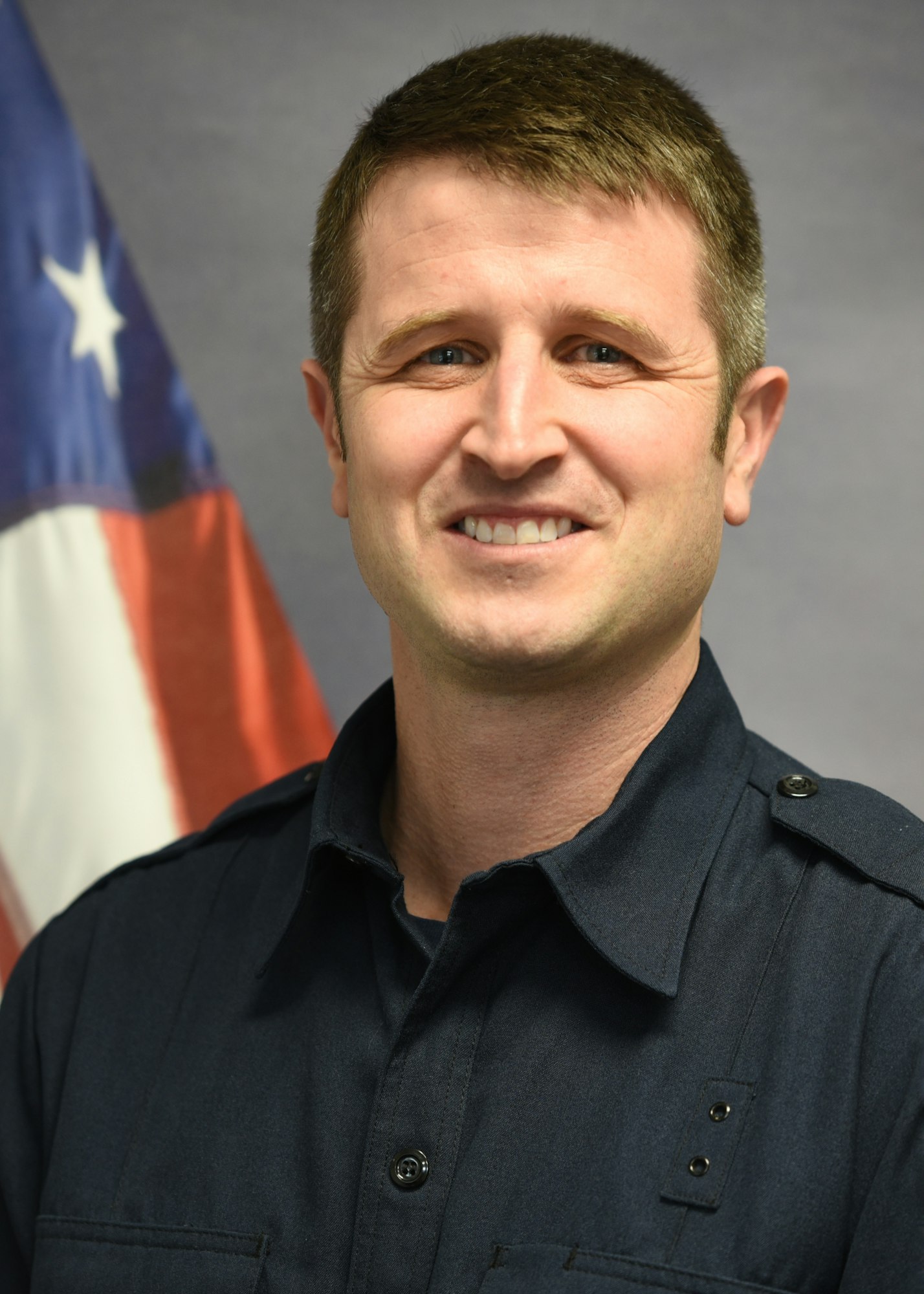 Man in a dark uniform standing in front of an American flag, smiling at the camera.