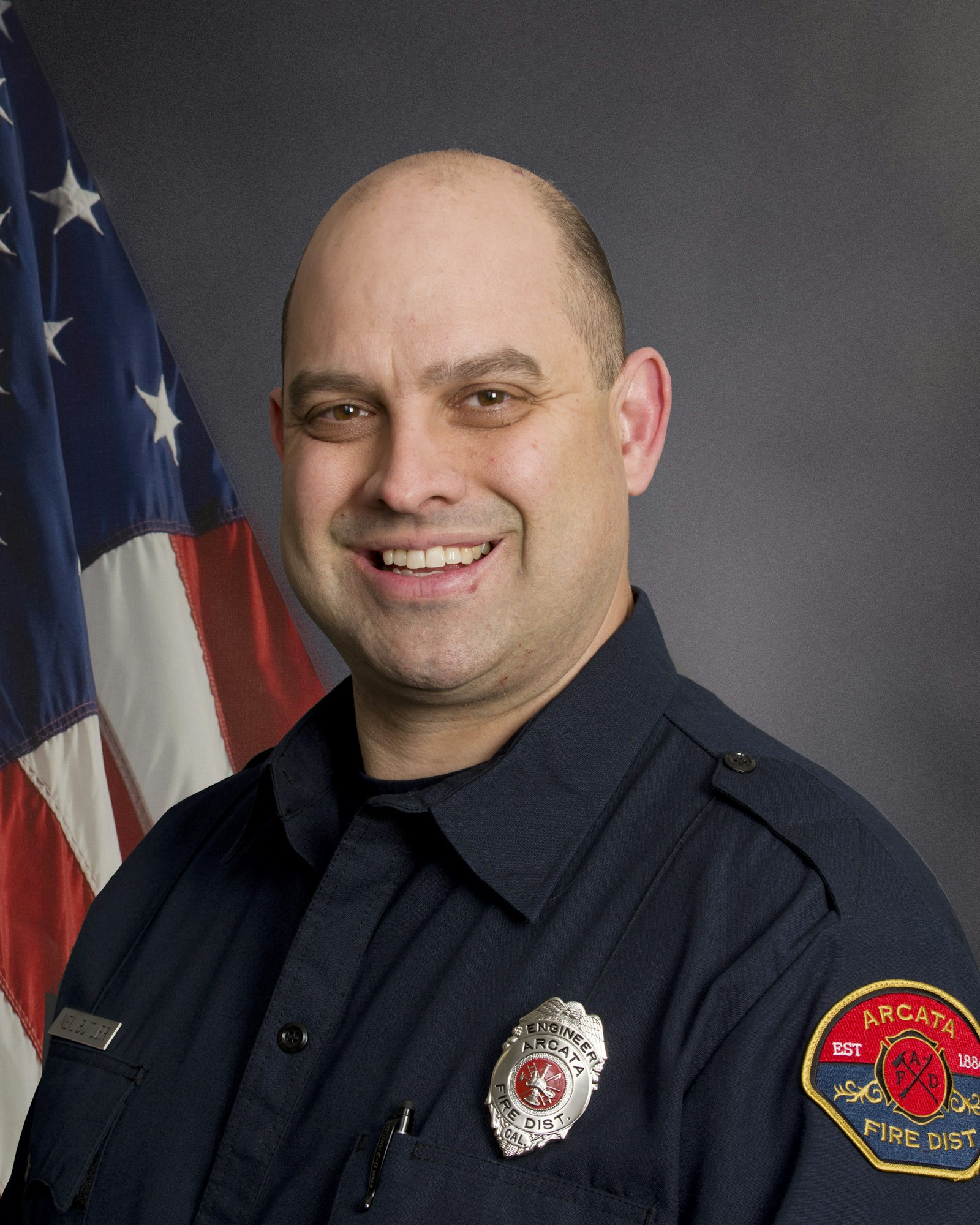 A smiling person in a firefighter uniform with badges, standing in front of an American flag.