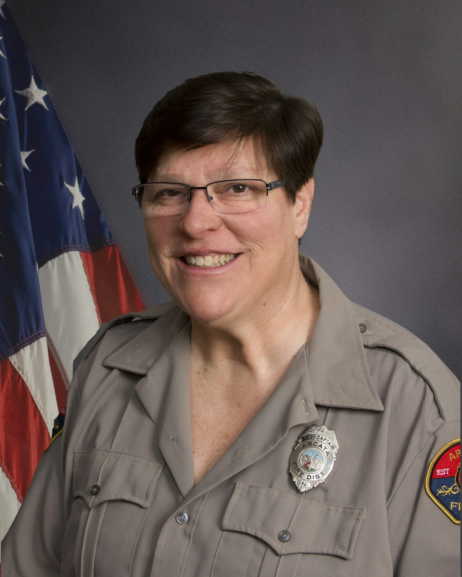 A person in a uniform with a badge, smiling, next to the American flag.