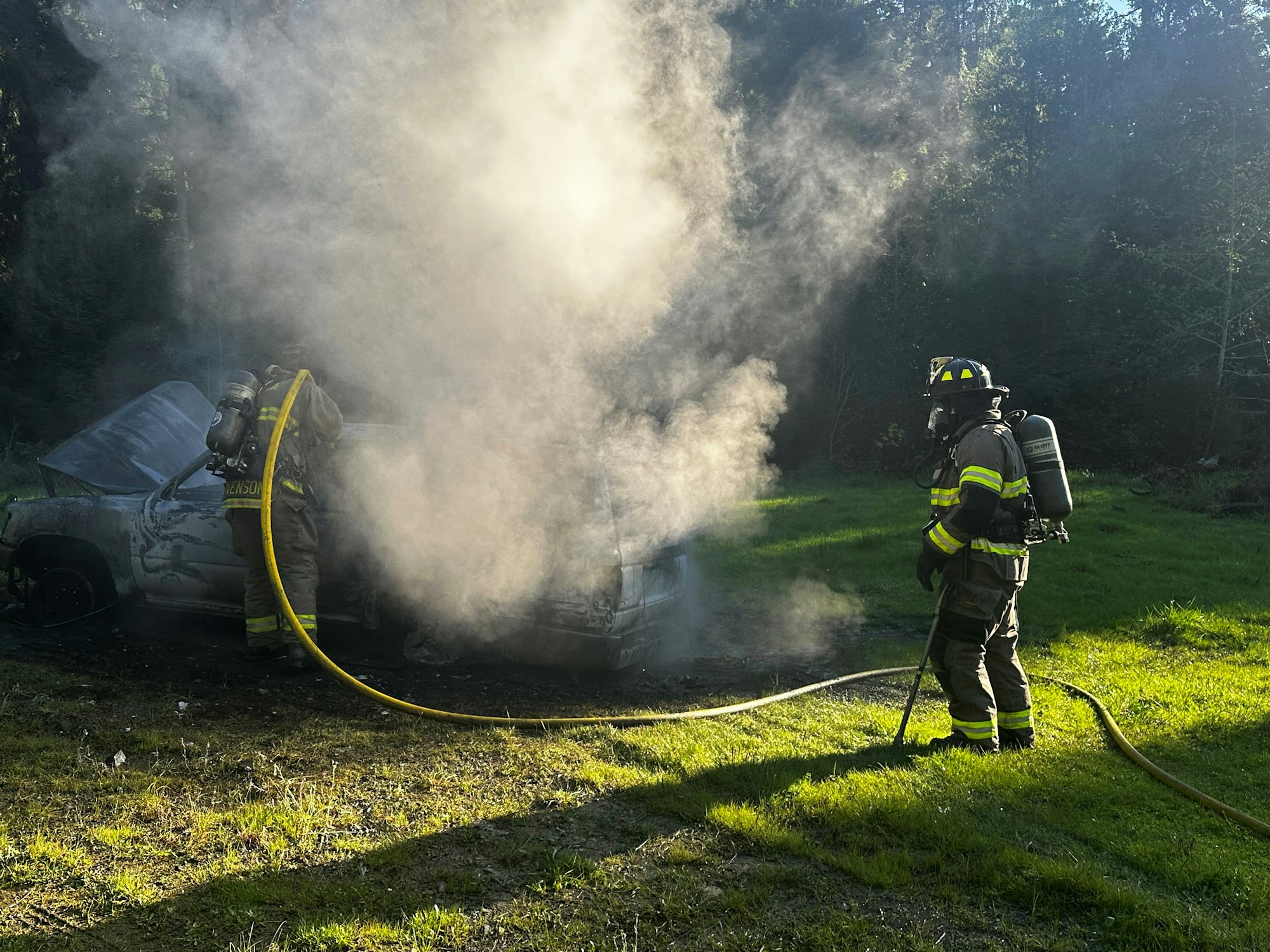 A firefighter extinguishing a smoking car in a grassy area.