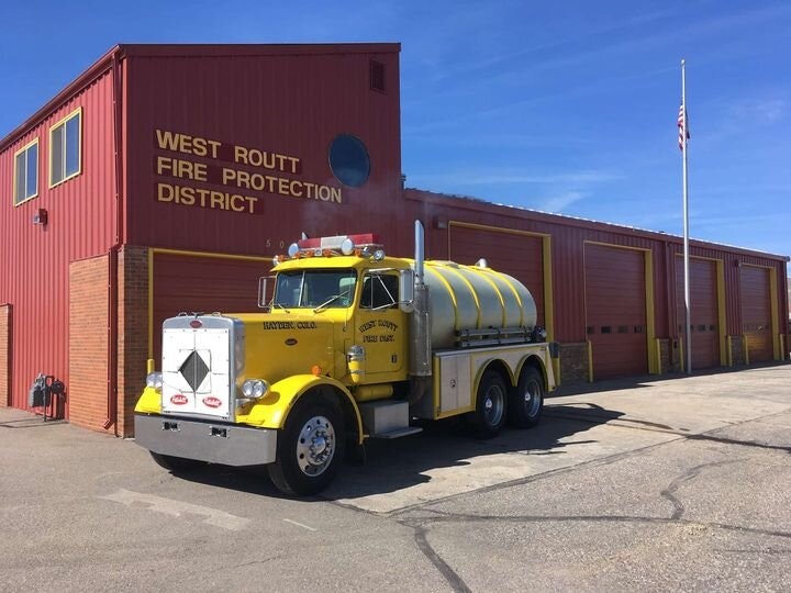 A yellow fire tanker truck parked in front of a fire station with the flagpole to the right.