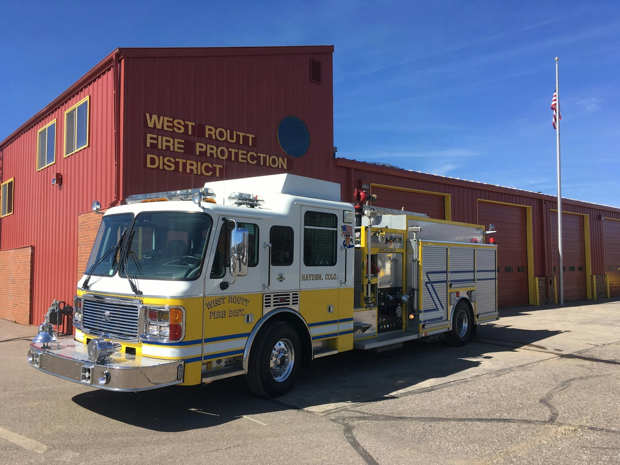 A fire truck parked outside a fire station with "West Routt Fire Protection District" signage and an American flag.