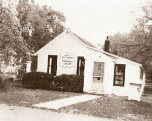 The image shows a small, quaint building identified as a library, surrounded by greenery. It's a simple, one-story structure.
