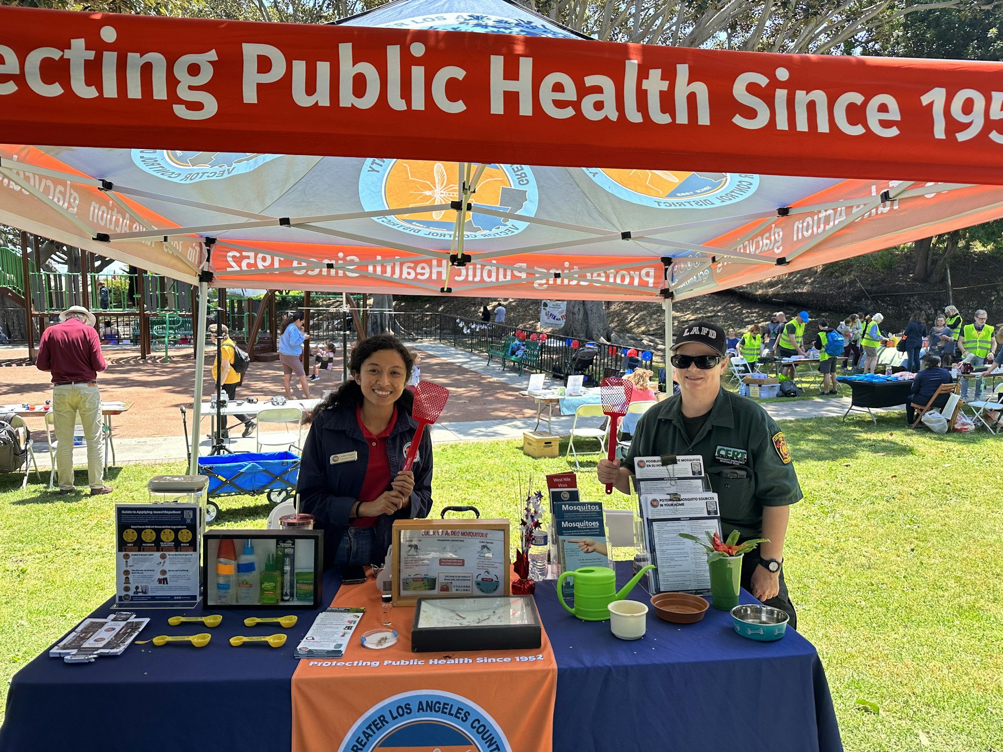A health event booth with two people displaying public health information and tools under a banner reading "Protecting Public Health Since 1958".