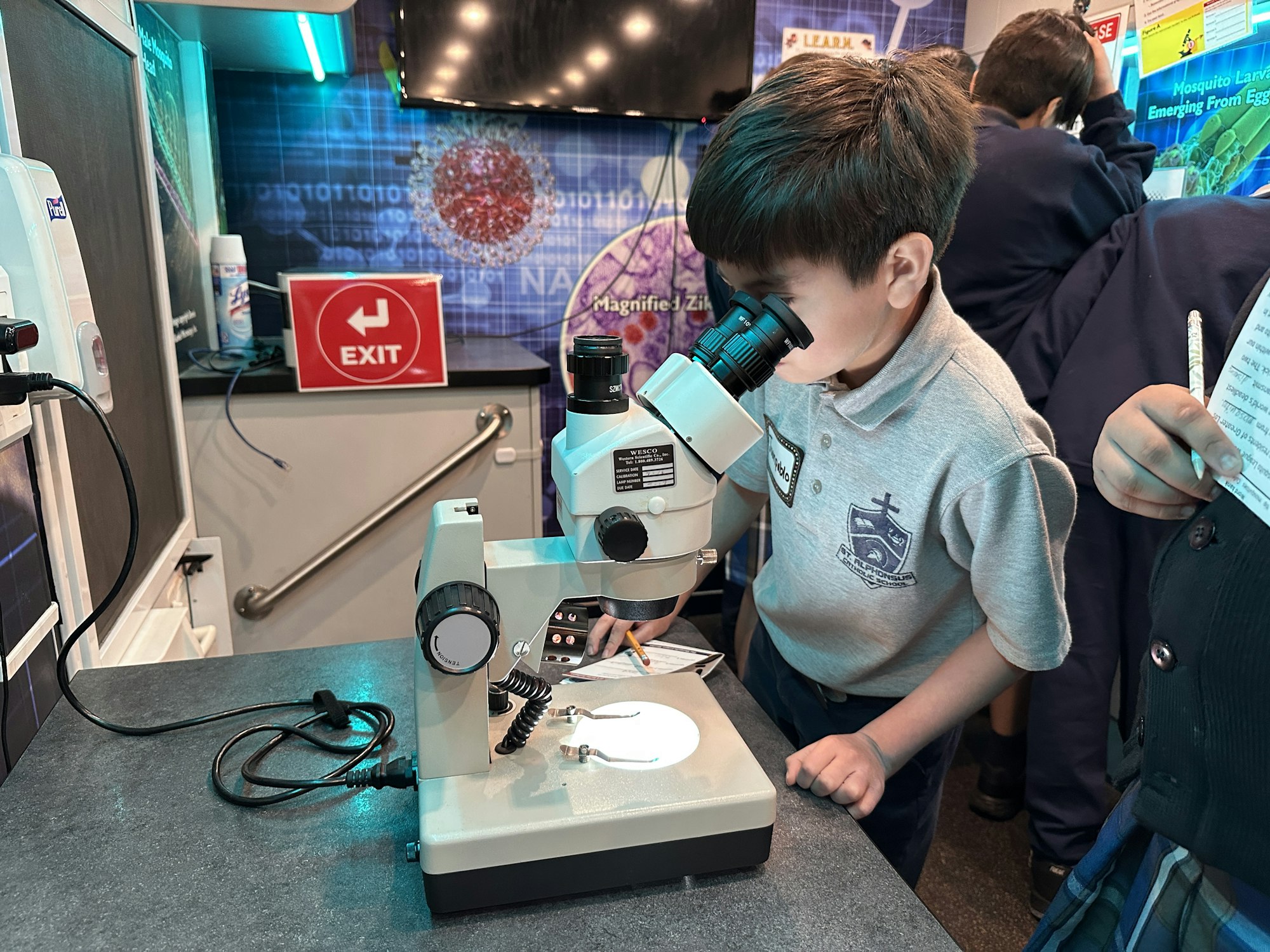 A child is looking through a microscope at a science exhibit with educational posters in the background.